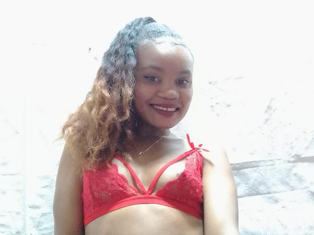 OdiJolie with curly brown hair, smiling, wearing a red lace bra, showing her small tits and cute face, standing in bright light.