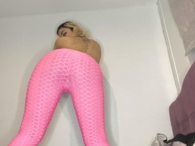 TarahDustin is topless, wearing pink, honeycomb-patterned leggings, with her back to the camera, looking over her shoulder. Her ass is prominent. She has blonde hair.