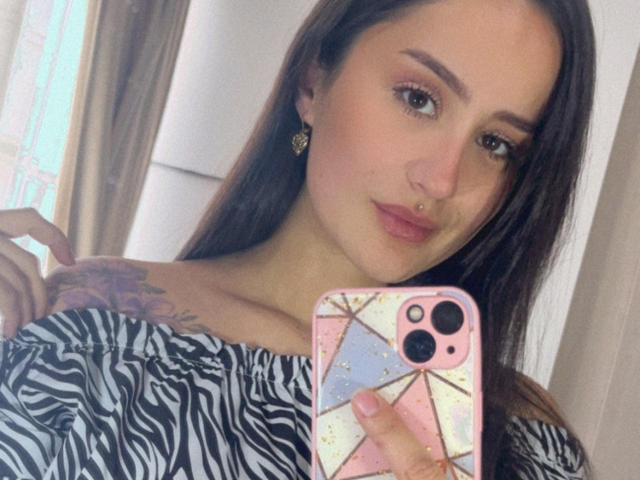 NinaBryons with long brown hair, zebra print top, pink phone case, nose ring, and tattoo on right shoulder, holding phone in front of mirror. She's looking at the camera.