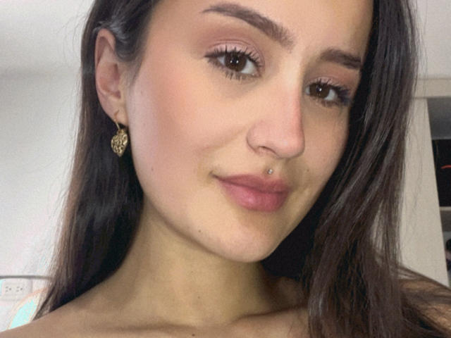 NinaBryons has long brown hair, brown eyes, a nose piercing, and a lip piercing. She's wearing gold heart earrings and smiling slightly. Her skin is light and smooth.