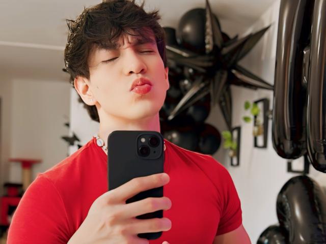 NeythanBlaze, with pouty red lips, short brown hair, and a red shirt, takes a selfie with a black phone, eyes closed, in a brightly lit room.