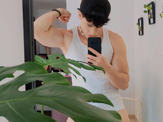 NeythanBlaze, muscular, flexing bicep, white tank top, black cap, gold chain, taking selfie, holding phone, green plant in foreground, white walls, black mirror frame, black wristband.
