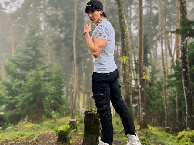NeythanBlaze standing in a forest, wearing a black cap, light gray shirt, black ripped jeans, and white sneakers, looking back over his shoulder with a slight smile.