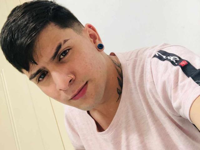 Sthefano has short black hair, blue ear gauges, a nose ring, and a tattoo on his neck. He's wearing a pink t-shirt with black stripes, looking at the camera.