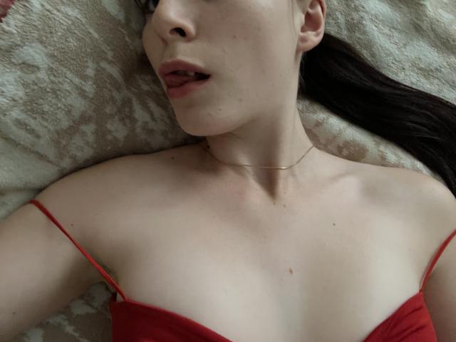 Valissias Gesicht und Brust, rotes Kleid mit Trägern, kleine Brüste, blasse Haut, braunes Haar, leicht geöffneter Mund, goldene Halskette, liegt auf einer gemusterten Decke, sichtbares Schlüsselbein, dezentes Make-up..