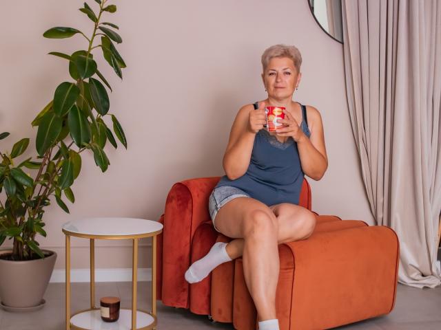EmiliaGree with short blonde hair, blue tank top, and denim shorts, sitting on an orange armchair, holding a red and white mug, wearing white socks, looking at camera.