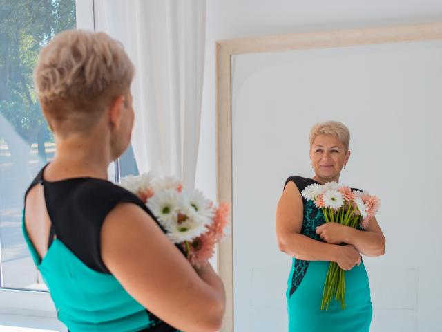 Emilia, con cabello rubio corto, un vestido verde azulado y negro, sosteniendo flores rosas y blancas, de pie frente a un espejo, que refleja su rostro sonriente, en una habitación luminosa..