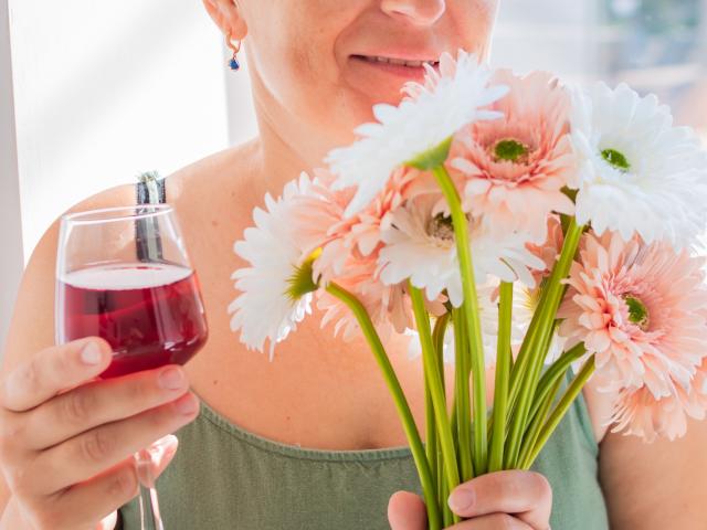 EmiliaGree holding a bouquet of pink and white gerbera daisies with her right hand, while holding a glass of red wine with her left hand.
