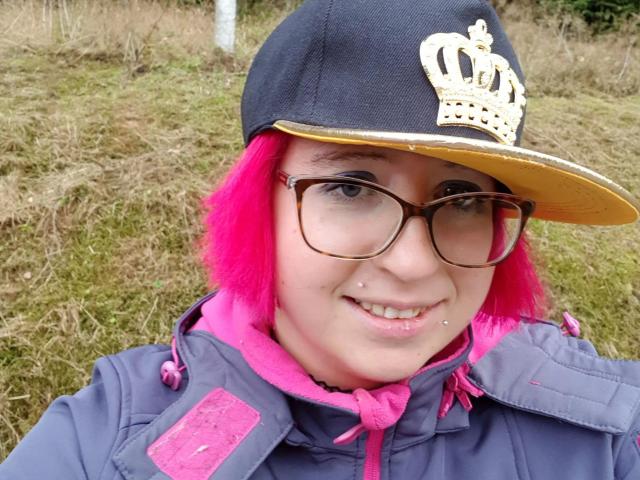Zaubermaus83-hot with bright pink hair, glasses, and a black baseball cap with a gold crown, wearing a purple jacket with pink trim, smiling with a nose piercing.