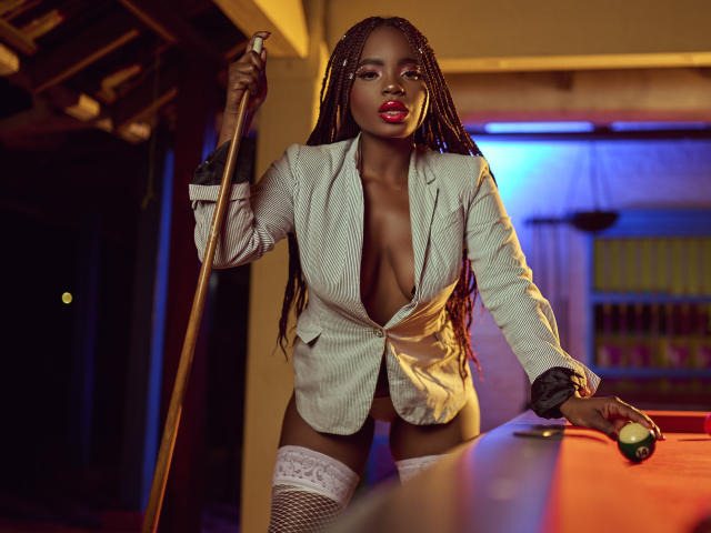 LindaBird with dark skin, braided hair, red lips, white blazer, deep cleavage, white lace stockings, holding pool cue, leaning on pool table, green ball in foreground.