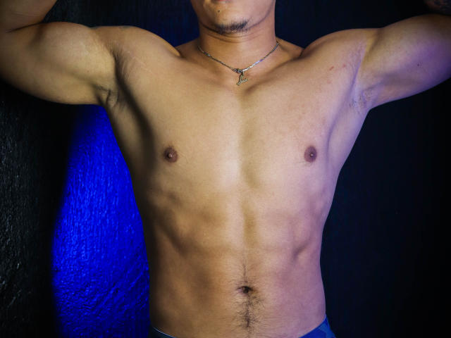 El cuerpo tonificado y sin camisa de JeisonStark, con abdominales visibles, pezones pequeños y un collar. Sus brazos están levantados, mostrando vello axilar. Lleva pantalones azules, con una luz azul detrás..