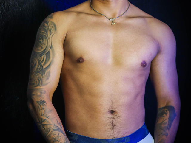 JeisonStark's shirtless upper body, showing tan skin, tattoos on both arms, a small necklace, and a slight belly with a visible navel. He's wearing white and blue pants.