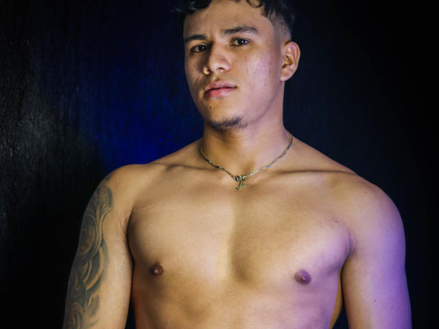 shirtless JeisonStark with a medium build, tattoos on the left arm, necklace, and a slight smirk, short curly hair, and a clean-shaven face. He's looking at the camera.