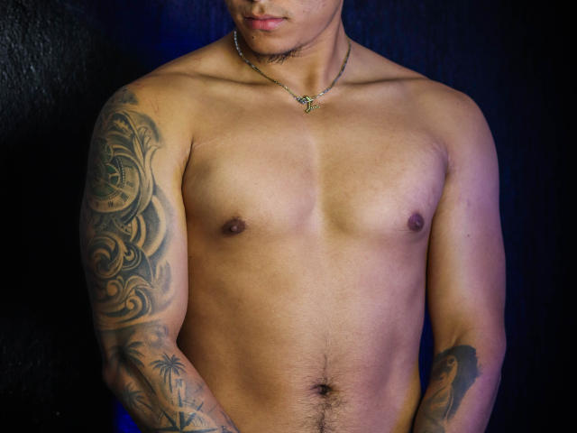 JeisonStark's shirtless upper body, showing a tattooed arm with swirls, a necklace with a small pendant, and a slight chest hair, against a dark background.