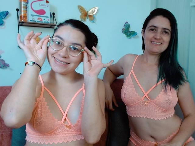 Two LunaVenus with dark hair, glasses, and pink lace bras with cross straps, smiling and adjusting glasses, showing their small tits and flat bellies, sitting on a couch.