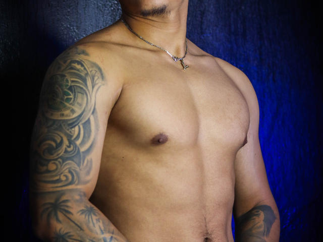 JeisonStark's shirtless torso, showing tattoos on both arms, small nipples, and a silver necklace with a small cross. His skin is tan, and he's standing against a dark background.