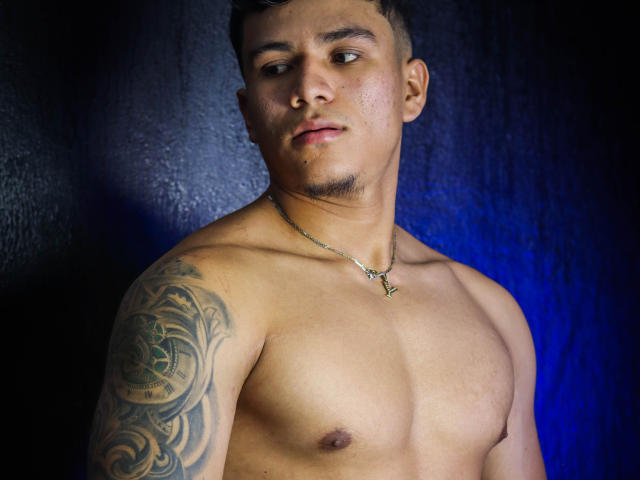 Shirtless JeisonStark with a tattoo on his right arm, wearing a gold necklace, has a serious expression, and short dark hair, against a dark blue background.