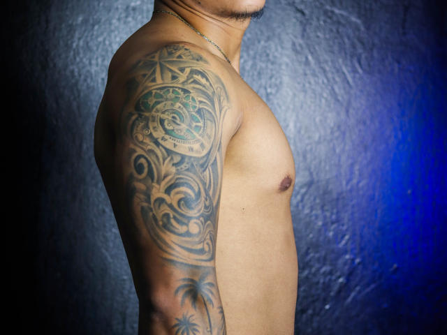 JeisonStark's tattooed arm, showing intricate, swirling patterns in dark green and black, with a small palm tree at the bottom, on his tan, shirtless, muscular upper body.