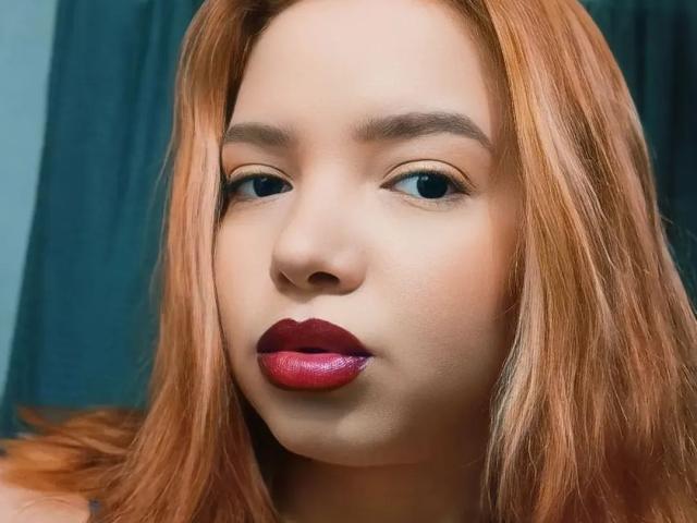 AuroraLiz has red lipstick, red hair, and blue eyes with light makeup. Her nose is centered, and she's looking slightly to the left. Her skin is fair.