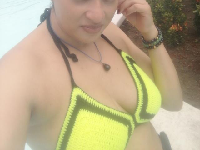 VioletaSwitch with yellow crochet bikini top, showing her big boobs, brown hair in braids, black necklace, and green bracelet, taking a selfie. Her skin is light brown.