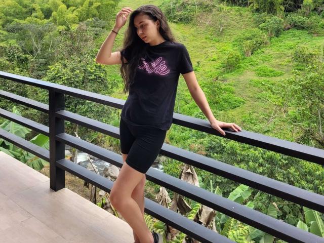 Emma Wells, com cabelos longos e escuros, vestindo uma camiseta preta e shorts pretos, apoiada em um corrimão preto, em pé em uma plataforma de azulejos, olhando para baixo, com a mão no cabelo..