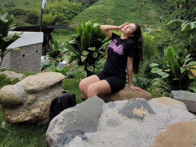 Emma Wells sentada em pedras, vestindo shorts pretos e uma camiseta rosa com estampa, com a mão no cabelo, sorrindo, com vegetação ao redor, uma mochila preta na grama, colinas exuberantes ao fundo..