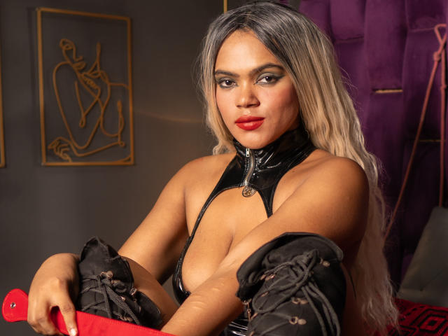 AliceJelanni with blonde hair, red lipstick, black leather top, and black boots, holding a red object, showing her tits, looking at the camera with a confident smile.