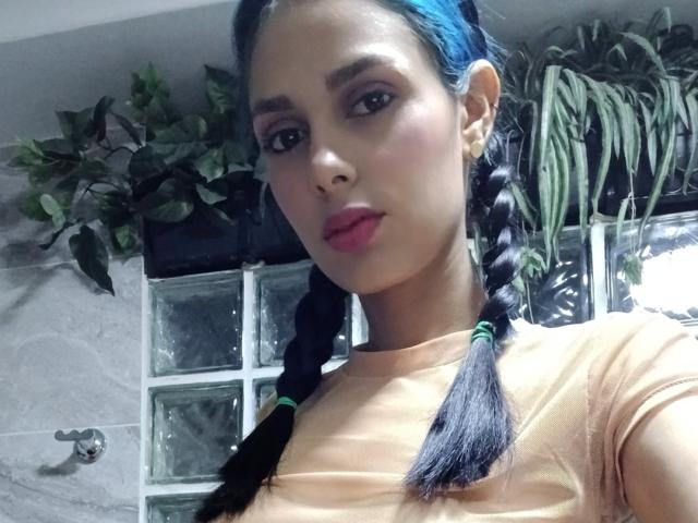 AnaisHilton with blue braided hair, tan shirt, pink lipstick, medium tits, brown skin, and green earbuds, in a bathroom with plants and glass blocks.