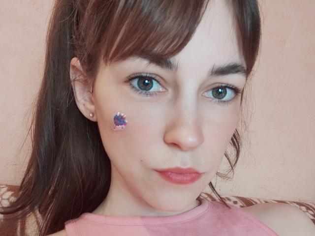 Barbariana with brown hair in a ponytail, green eyes, pink lips, and a purple butterfly sticker on her cheek, wearing a pink shirt, pouty expression.
