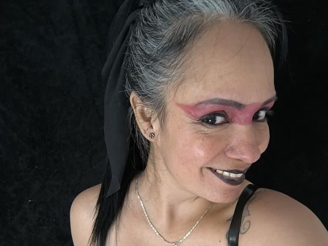 LailaCastillo with gray hair, black lipstick, pink eyeshadow, black ribbon, smiling, showing off her tits, black strap, small earrings, and a silver necklace against a dark background.