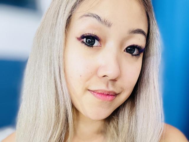 RenyLime has light blonde hair, purple eyeshadow, long eyelashes, and pink lipstick, with a small beauty mark on her left cheek, smiling softly at the camera.