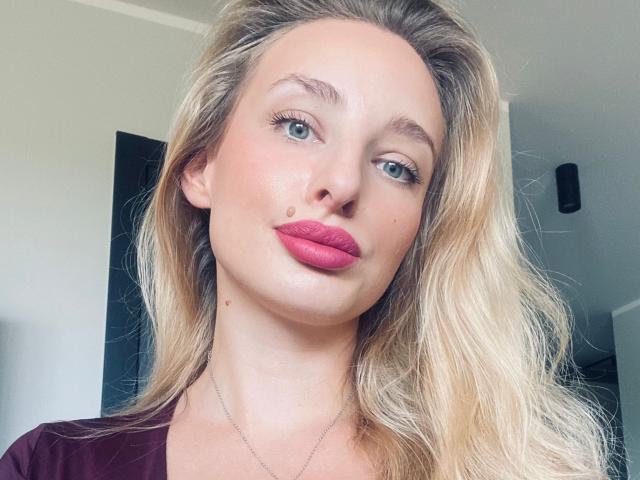 MarilynPretty tem cabelo loiro, olhos azuis, batom rosa e uma pequena marca de beleza na bochecha direita. Ela está vestindo uma blusa marrom e um colar fino..