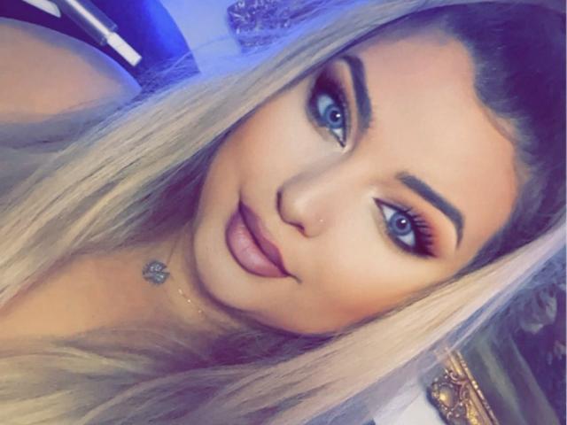 AllysonK has long blonde hair, blue eyes, and full pink lips with a subtle smile. She's wearing dark eyeliner and a small silver necklace with a flower.