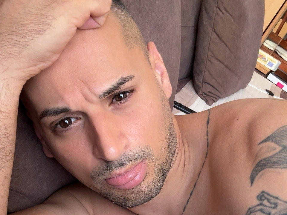 LexJay69, shirtless with a shaved head, brown eyes, and a tattoo on his right shoulder, looks slightly confused with a hand on his head, pouty pink lips, and a light beard.