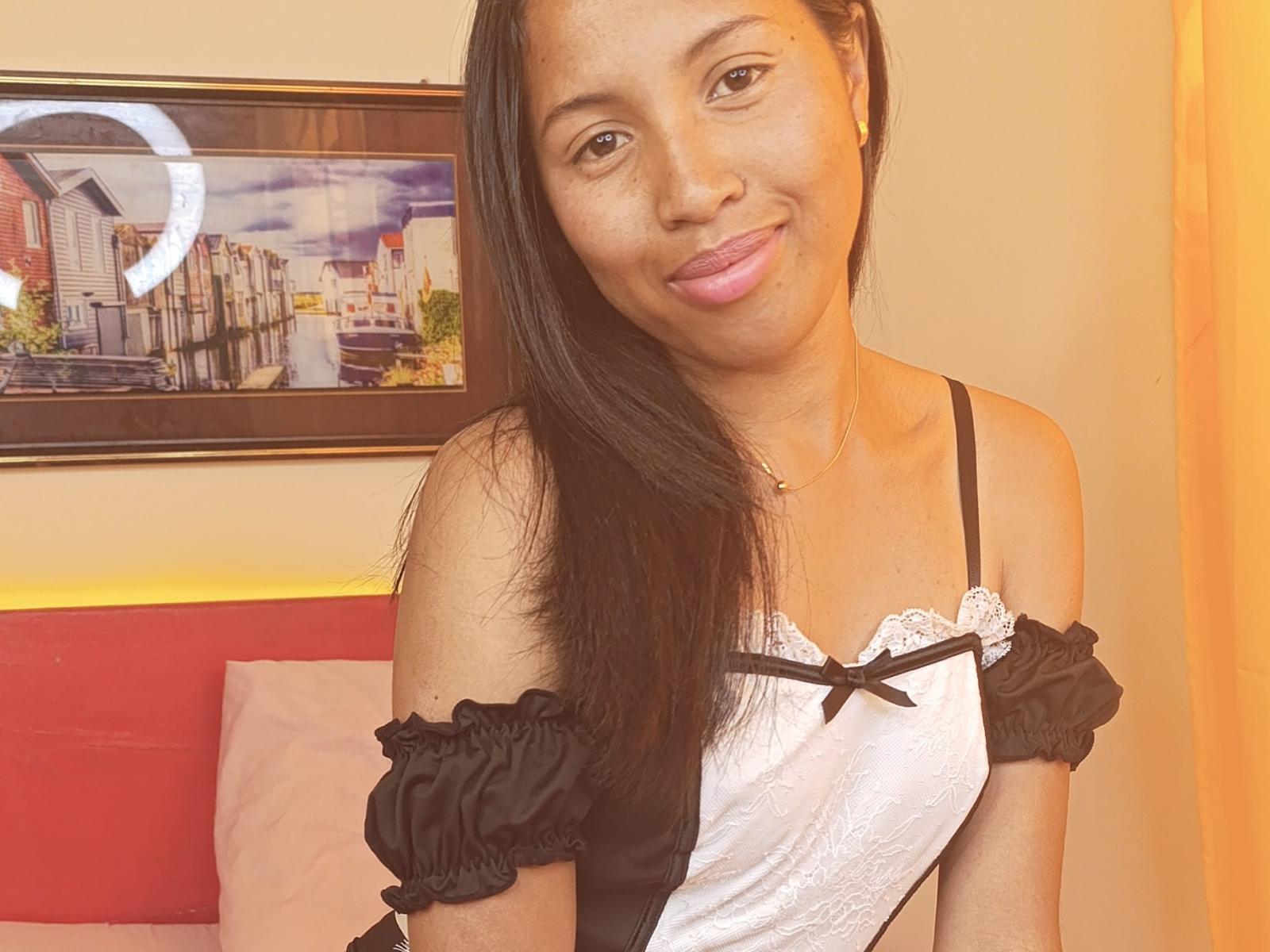 CristalDesiree, com longos cabelos pretos, sorrindo, vestindo uma blusa preta e branca com um ombro de fora, mostrando seus pequenos seios, em um quarto aconchegante com uma foto da cidade emoldurada..