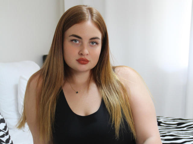 EsterMiln has long brown hair, blue eyes, and wears a black tank top, sitting with a neutral expression and red lipstick, showing her small tits and fair skin.