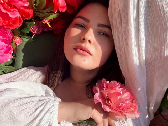 Bluria con un fiore rosa in mano, sdraiata, vestito bianco, viso illuminato dal sole, capelli castani, rose rosa, foglie verdi, espressione dolce, luce naturale, sfondo floreale, ombre delicate, rilassata..
