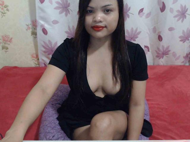 CreamyCarmen with medium tits in a deep black dress, sitting on a red and purple bed, long brown hair, red lipstick, smiling, arm resting on bed, background curtain.