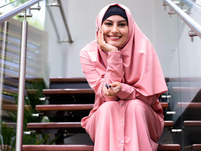 SajarBentaleb in pink outfit, black hijab, smiling, crouched on stairs, holding black pen, hands on cheek, metal railing, wooden steps, glass panels, green plant in background.