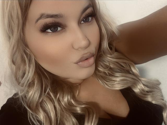 AllysonK with blonde wavy hair, full eyebrows, long eyelashes, and pouty pink lips, wearing a black top, showing her cleavage and slightly tanned skin.