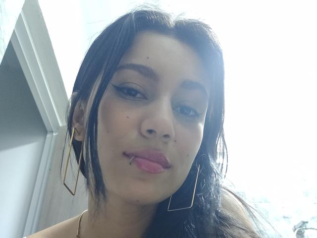 KataleyaMansson has dark hair, pouty pink lips, light makeup, gold earrings, and a small mole on her right cheek. Bright sunlight highlights her face.