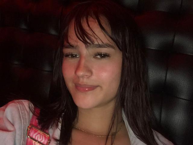 MelodyMansson has wet brown hair, light skin, and a subtle smile, wearing a pink jacket with a gold necklace, in a dark leather booth.