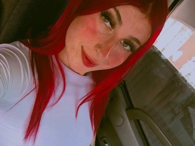 SammyViolet with red hair, wearing a white turtleneck, has her tits pushed up, a nose ring, and is smiling in a car, looking at the camera.