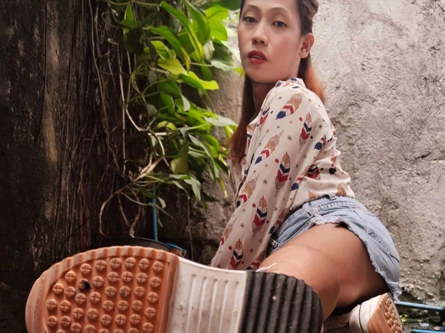 Darcy, no verão, ajoelhada, exibindo o bumbum em shorts jeans rasgados, vestindo uma blusa estampada, com o rosto voltado para a câmera, e a sola do sapato em primeiro plano..