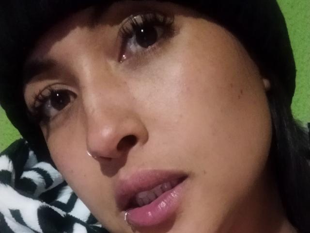 O rosto de MirandaDreams, olhos castanhos, cílios longos, piercing no nariz, gorro preto, piercing rosa nos lábios, lábios rosa ligeiramente entreabertos, fundo verde, tecido preto e branco estampado no canto inferior esquerdo..