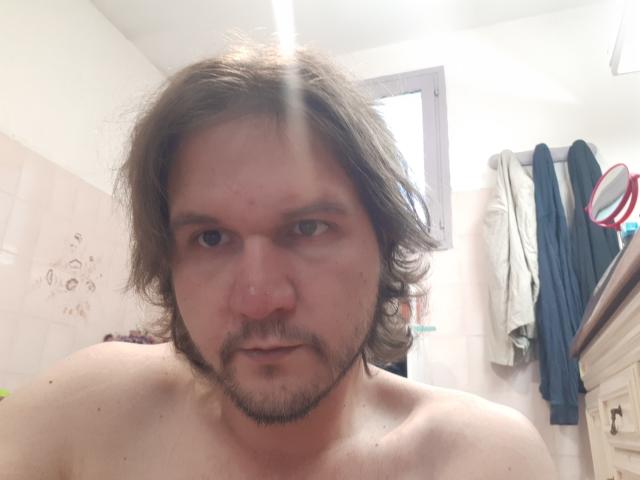 NectarineFad tem uma constituição média, cabelo castanho ondulado e barba. Ele está sem camisa, mostrando um nariz levemente avermelhado e uma expressão séria. A luz do sol brilha em seu rosto..