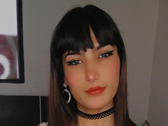 Lunardy has black eyeliner, blue eyes, a nose ring, and a choker. She's wearing red lipstick and has straight brown hair with bangs. She's also got hoop earrings.