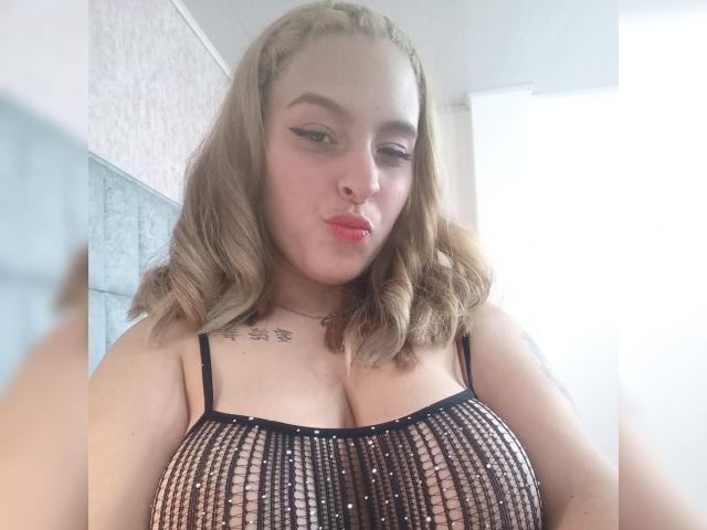StacyAnzola with blonde wavy hair, pouty red lips, wearing a black beaded mesh top, showing her big tits, tattoo on chest, and a nose ring, looking down.