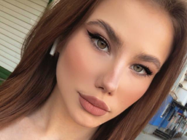 NicoleNight has green eyes, brown eyeliner, and brown lipstick. Her red hair is down, and she's wearing white earbuds. Her face is slightly flushed, looking at the camera.