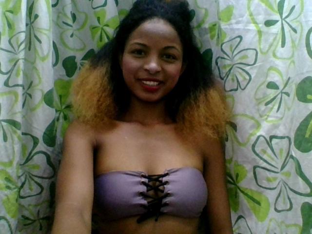 AlexaChan with curly black and blonde hair, smiling, wearing a purple lace-up bralette, showing her tits, standing in front of a green and white floral curtain.
